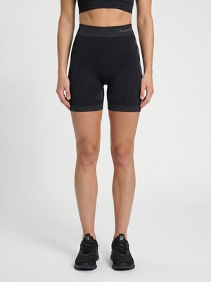 Femme portant un short de sport noir taille haute et des chaussures de course noires, debout devant un fond clair uni.