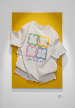 White short-sleeve shirt displayed on a yellow background, featuring a colorful four-square graphic with flower shapes and text below.