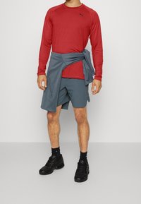 Sportoutfit bestehend aus einem roten Langarmshirt, grauen Shorts und schwarzen Turnschuhen. Das Shirt hat ein Logo auf der Brust; die Shorts haben Taschen.