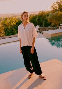 Γυναίκα με λευκό πουκάμισο κοντομάνικο και μαύρο φαρδύ παντελόνι, στέκεται δίπλα σε μια πισίνα τη δύση του ηλίου με δέντρα και ξαπλώστρες στο φόντο.