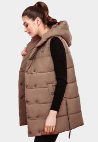 Beige Steppweste mit Kapuze, ausgestattet mit Druckknöpfen und einem seitlichen Reißverschluss. Strukturierter Stoff mit gesteppte Abschnitten, getragen über einem schwarzen Rippenshirt.