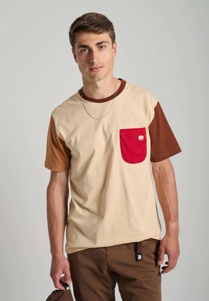 Junger Mann trägt beige-braunes T-Shirt mit roter Tasche und braunen Hosen, steht vor neutralem grauem Hintergrund.