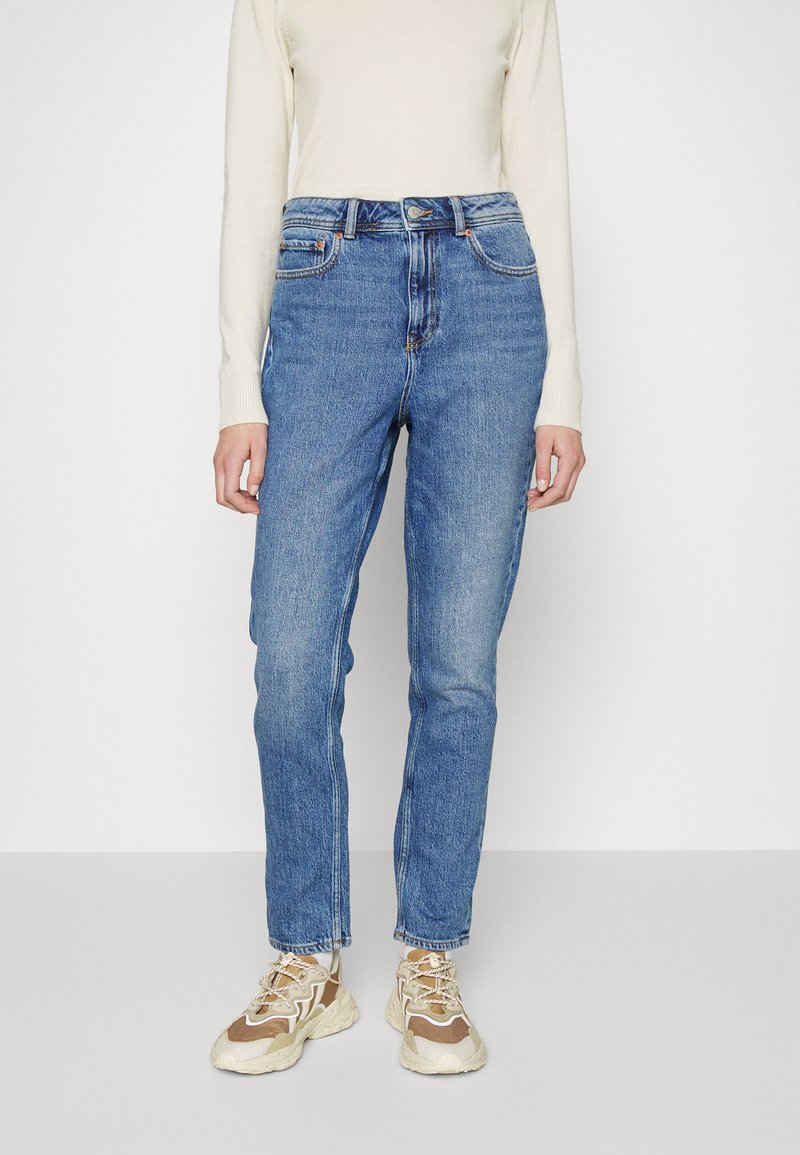 Jeans de cintura alta azul-claro com cinco bolsos, fecho de botão e zíper, e um design de perna reta, usados com ténis bege.