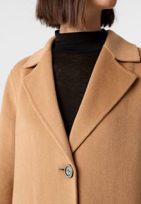 Donna che indossa un cappotto di lana color cammello sopra un dolcevita nero, mostrata dalle spalle fino a metà torso, con un singolo bottone allacciato.