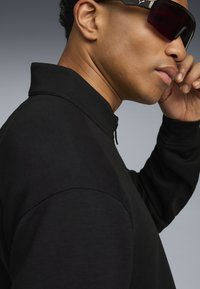 Mand, der justerer sorte sportssolbriller, iført sort zip-up trøje med høj krave, mod en neutral grå baggrund.