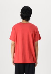 Tricou roșu din bumbac, cu mâneci scurte și guler rotund, având o croială lejeră și o textură netedă, cu cusături vizibile la tiv.