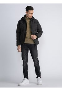 Chaqueta de plumas negra con capucha, cierre de cremallera, llevada sobre un suéter verde. Combinada con jeans oscuros y zapatillas blancas. Ajuste casual.