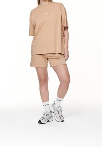 Donna che indossa una t-shirt beige oversize e pantaloni corti, con calze bianche e sneakers grigie e bianche, in piedi con una gamba leggermente piegata.
