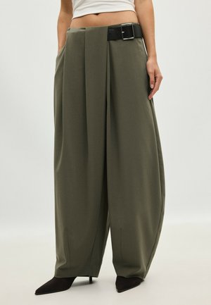 Personne portant un pantalon large vert olive avec des plis, une ceinture noire, des talons pointus noirs et un top court blanc, debout sur un fond clair.