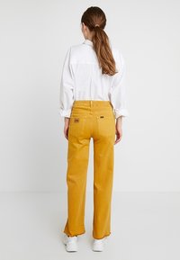 Pantalones de denim amarillos de pierna ancha, con una cintura alta, dos bolsillos traseros y un dobladillo deshilachado, combinados con una camisa blanca de manga larga.