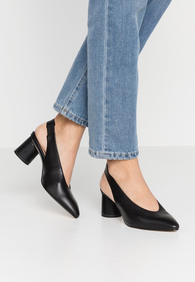 Escarpins à talons carrés en cuir noir avec bride arrière portés avec un jean bleu clair sur une personne debout sur un sol blanc.