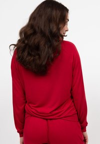 Rode langhouder top gemaakt van zachte stof, met een losse pasvorm en een gebogen zoom. Draagt met bijpassende broeken, wat een ontspannen silhouet laat zien.