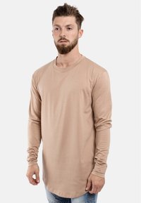 Cămașă bej cu mânecă lungă dintr-un material neted, cu guler rotund și tiv curbat, purtată cu blugi albastre. Design simplu, fără modele vizibile.