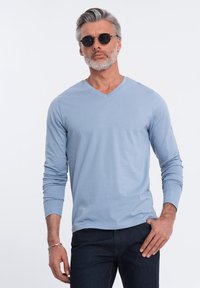 Hellblaues Langarmshirt mit V-Ausschnitt aus weichem Stoff, kombiniert mit dunklen Jeans. Mann trägt runde schwarze Sonnenbrille.