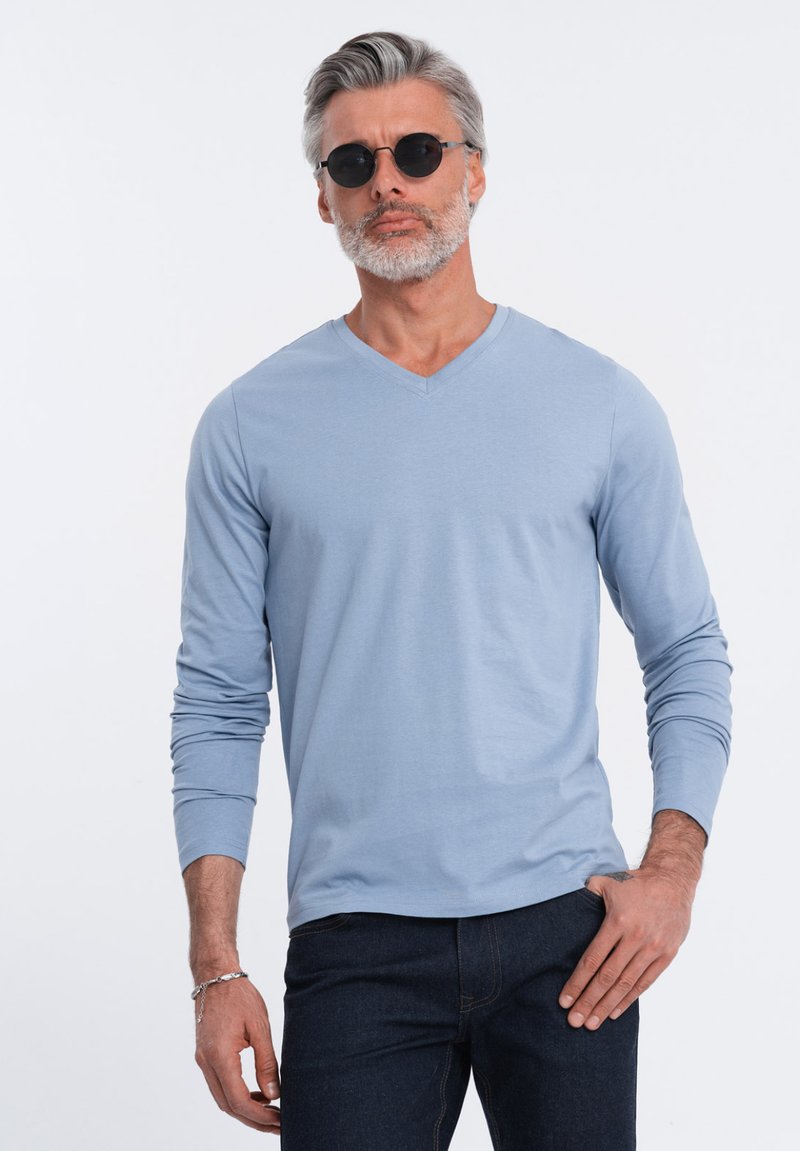 Hellblaues Langarmshirt mit V-Ausschnitt aus weichem Stoff, kombiniert mit dunklen Jeans. Mann trägt runde schwarze Sonnenbrille.