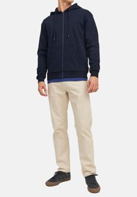 Marineblauwe zip-up hoodie met verstelbare trekkoord, kangoeroezakken, gedragen over een blauwe shirt, gecombineerd met lichtbeige broek en zwarte sneakers.
