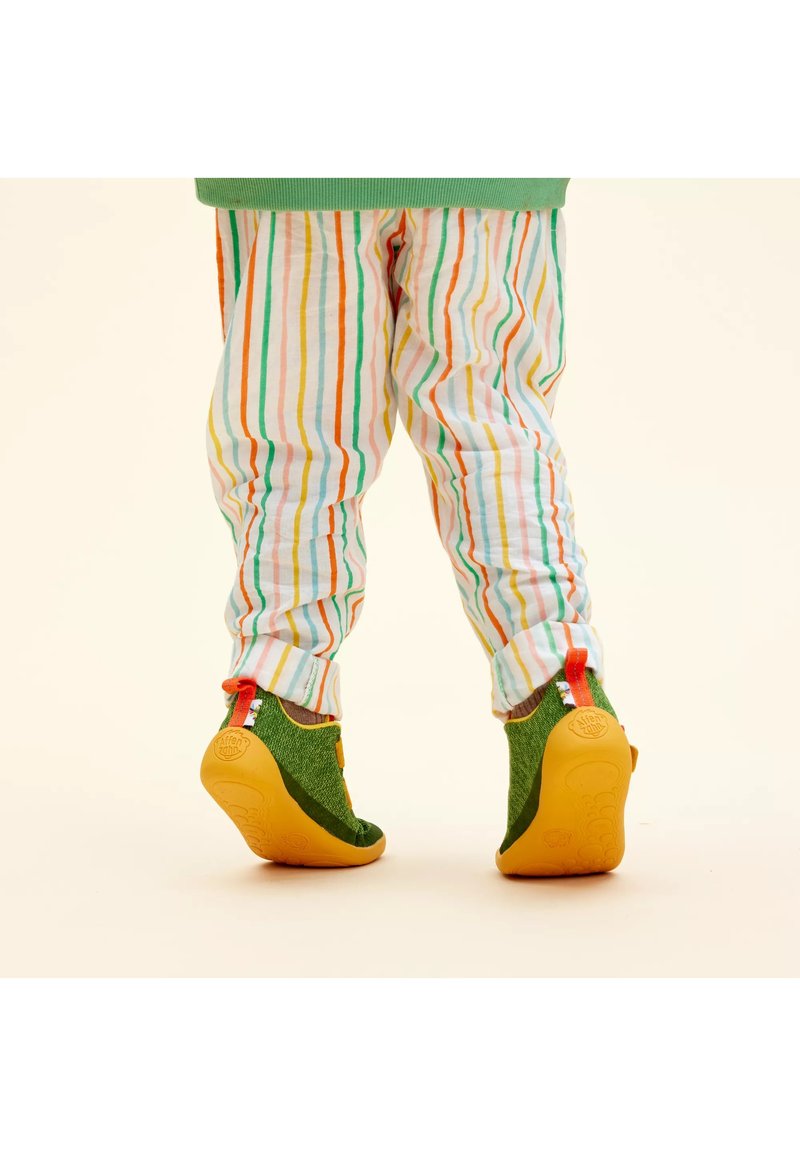 Kind trägt mehrfarbige gestreifte Hose und grüne Schuhe mit gelben Sohlen, steht auf Zehenspitzen vor einem einfarbigen Hintergrund.