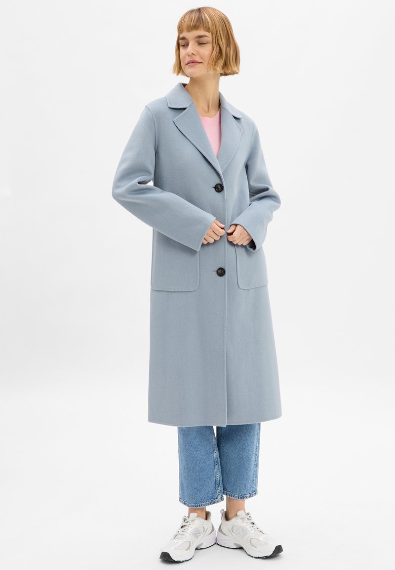 Cappotto lungo azzurro chiaro con colletto a punte, chiusura con bottoni e tasche laterali. Abbinato a un top rosa e jeans cropped, indossato con scarpe sportive.
