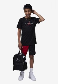 Berniukas, vilkintis juodai raudonais Jordan šortais ir juodu Jordan marškinėliais, laiko juodą Jordan kuprinę su baltu logotipu, šypsosi ir reguliuoja juodą kepurę.