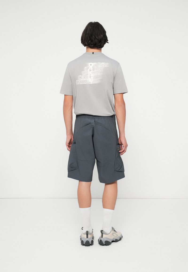 Graues T-Shirt mit bedrucktem Text auf dem Rücken, kombiniert mit dunkelgrauen Cargo-Shorts. Die Sneakers haben eine dicke Sohle und weiße Socken sind sichtbar.