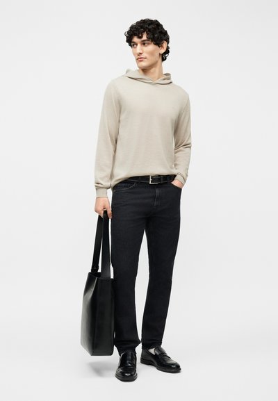 Νεαρός άνδρας με μπεζ πουλόβερ με κουκούλα, μαύρο τζιν, μαύρα δερμάτινα παπούτσια και κρατώντας μια μεγάλη μαύρη τσάντα ώμου, στέκεται μπροστά σε λευκό φόντο.