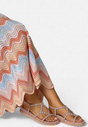 Piernas que llevan sandalias planas de tiras con cuentas rosas y una falda de punto fluida con rayas onduladas en tonos durazno, azul y beige.