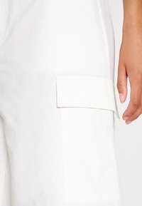 Witte cargobroek met een zijzak en zichtbare stiksels. Het materiaal lijkt lichtgewicht; een hand rust op de stof.