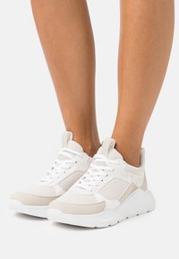 Zapatillas de deporte beige claro con acentos blancos, material de malla y sintético, detalles texturizados, cordones planos y una suela gruesa y texturizada.