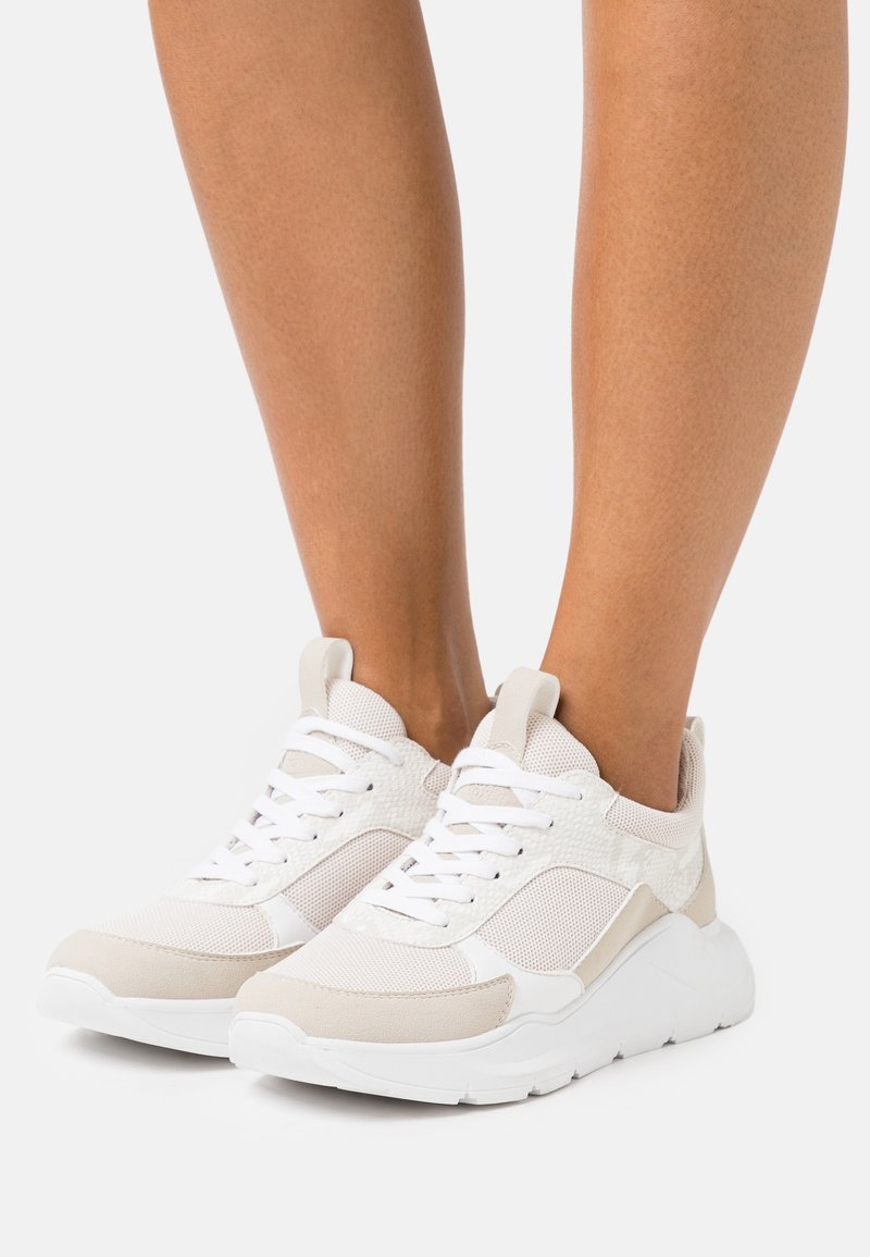 Zapatillas de deporte beige claro con acentos blancos, material de malla y sintético, detalles texturizados, cordones planos y una suela gruesa y texturizada.