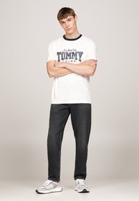 Maglietta bianca a maniche corte con colletto blu navy e grafica "TOMMY". Abbinata a jeans grigio scuro e sneaker grigie. Design semplice e vestibilità casual.