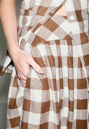 Main dans la poche d'une robe à carreaux marron et blanc avec une jupe plissée et un top sans manches assorti avec un bouton recouvert de tissu.