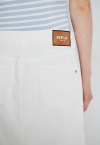 Hvit denimskjørt med en brun skinnlapp merket "REPLAY" og rød brodering på baklommen. Materialet er glatt, med et avslappet design.