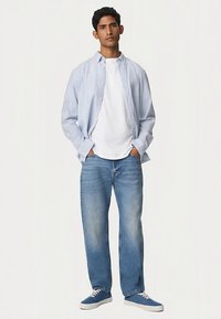 Lichtblauw-wit gestreept overhemd over een wit T-shirt, gecombineerd met mediumblauwe jeans en blauwe sneakers. Model staat met handen in de zakken.