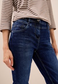 Jeans de mezclilla en azul oscuro con un ligero desvanecimiento, que cuentan con cinco bolsillos, acentos de costura y un cierre de botón. Camisa de manga larga a rayas cercana.