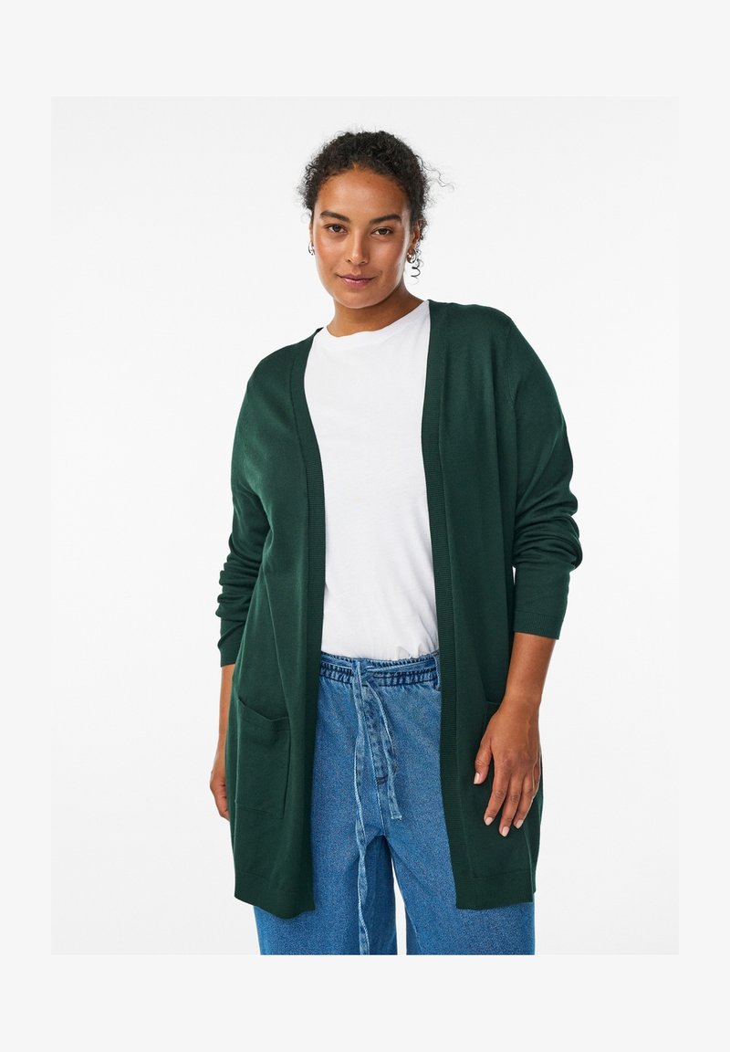Kvinde iført en mørkegrøn cardigan med åben front over en hvid t-shirt og løstsiddende blå jeans med knyttet taljebånd, stående mod en hvid baggrund.