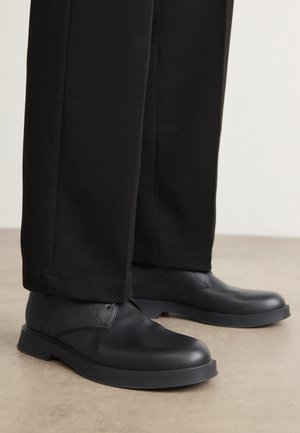 Mustad mustad nahast kingad ümarate varvastega, lame tallaga ja minimaalsete detailidega. Kombineeritud pritsitud mustade pükstega, mis näitavad siledat tekstuuri.