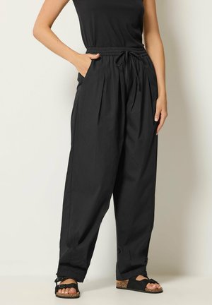 Vrouw draagt een losse zwarte broek met trekkoord in de taille, handen in de zakken, een zwarte mouwloze top en zwarte sandalen op een effen achtergrond.