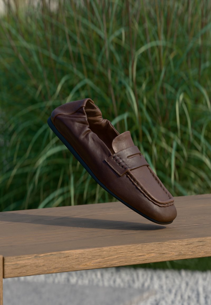 Mocassin en cuir marron flottant au-dessus d’un banc en bois avec de hautes herbes vertes floues en arrière-plan.