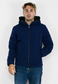 Giacca con zip di colore blu navy, dotata di cappuccio, con un esterno liscio, design imbottito e rivestimento interno verde a contrasto.
