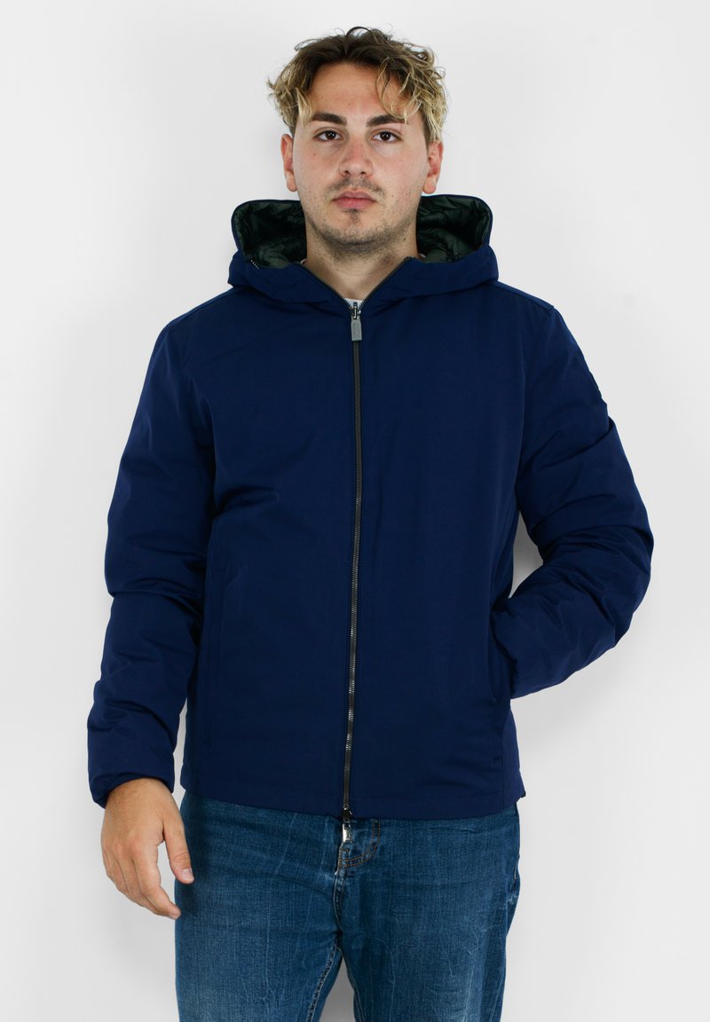 Giacca con zip di colore blu navy, dotata di cappuccio, con un esterno liscio, design imbottito e rivestimento interno verde a contrasto.
