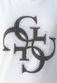 Camiseta blanca con un logotipo oscuro embellecido hecho de tachuelas redondas, con tachuelas plateadas más pequeñas esparcidas por el diseño.