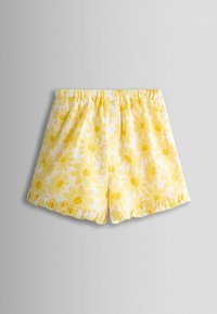 Pantaloni scurți cu imprimeu floral galben, cu talie elastică și tivuri cu volane pe un fundal simplu gri deschis.