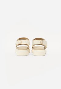 Beige sandaler med justerbare stropper, en teksturert overdel og en kraftig såle med et logo-emblem på baksiden.