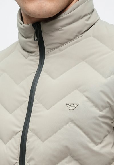 Világos bézs puffert kabát halszálka steppelt mintával, magas gallérral és fekete cipzárral. Kis logó található a mellrészen. Simább tapintású anyag.