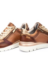 Baskets en cuir marron et beige avec un design texturé, des accents métalliques et une semelle extérieure blanche. Présente des lacets et des zips sur les côtés pour un accès facile.