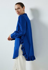 Cămașă albastru regal cu mâneci lungi, cu accente volăcelate pe laturi și la manșete, purtată cu pantaloni albi. Textură de țesătură netedă.