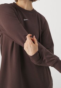 Persona che indossa una felpa Nike marrone scuro, mentre aggiusta o raddrizza la manica con una mano davanti a uno sfondo chiaro e semplice.