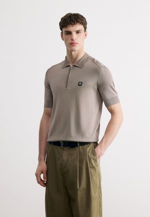 Ung mand iført en taupe kortærmet polo med lynlås og olivengrønne bukser, stående med den ene hånd i lommen mod en ensfarvet baggrund.