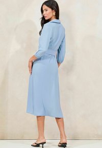 Abito azzurro chiaro, di lunghezza media, con colletto, maniche a sbuffo e vita delineata da una cintura. Tessuto liscio con silhouette fluida. Sandali neri con tacco.