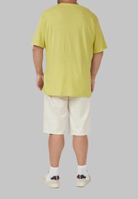 Żółta, fakturowana koszulka z krótkim rękawem, jasnobeżowe szorty i białe sneakersy z akcentami. Widok z tyłu na szarym tle.